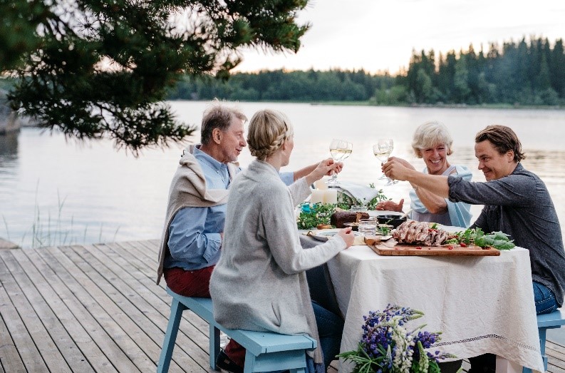 Hymyilevä pöytäseurue kohottaa kuohuviinilaseja järvimaisemassa.