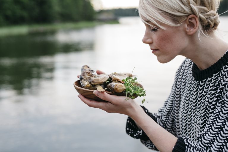 hymyilevä nainen katsoo savustettuja kaloja tarjottimella järvimaisemassa.