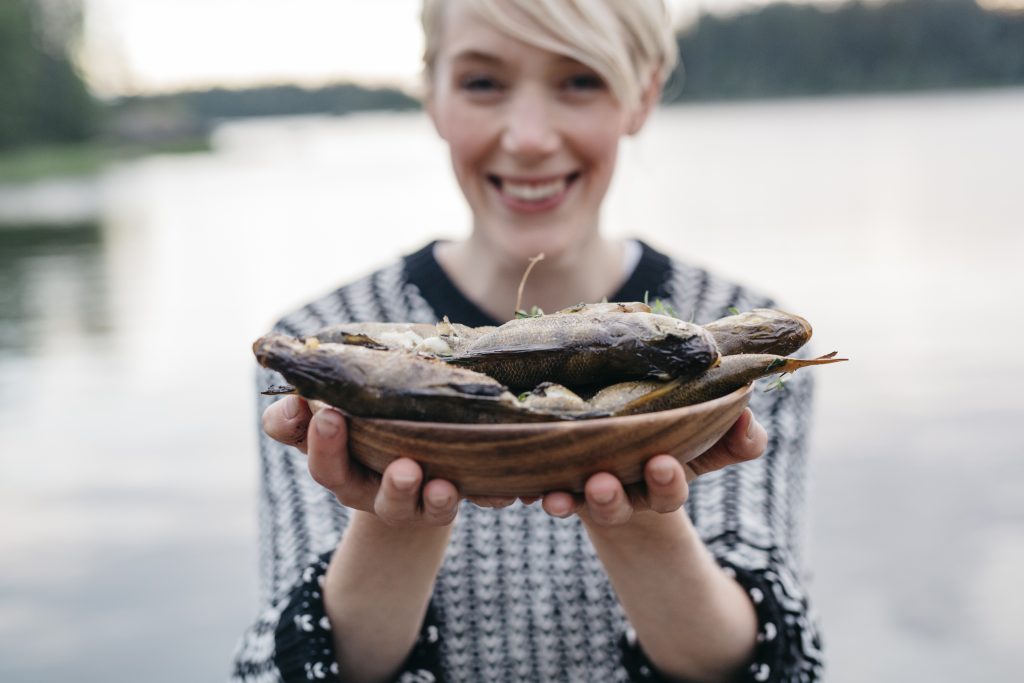 hymyilevä nainen ojentaa savustettuja kaloja kameraa kohti järvimaisemassa.