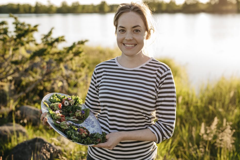 Nainen hymyilee ja pitelee tarjotinta täynnä kasvisannoksia järvimaisemassa.