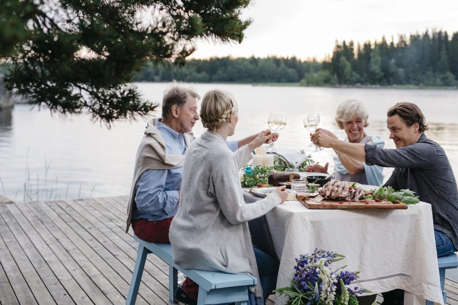 Hymyilevä pöytäseurue kohottaa kuohuviinilaseja järvimaisemassa.