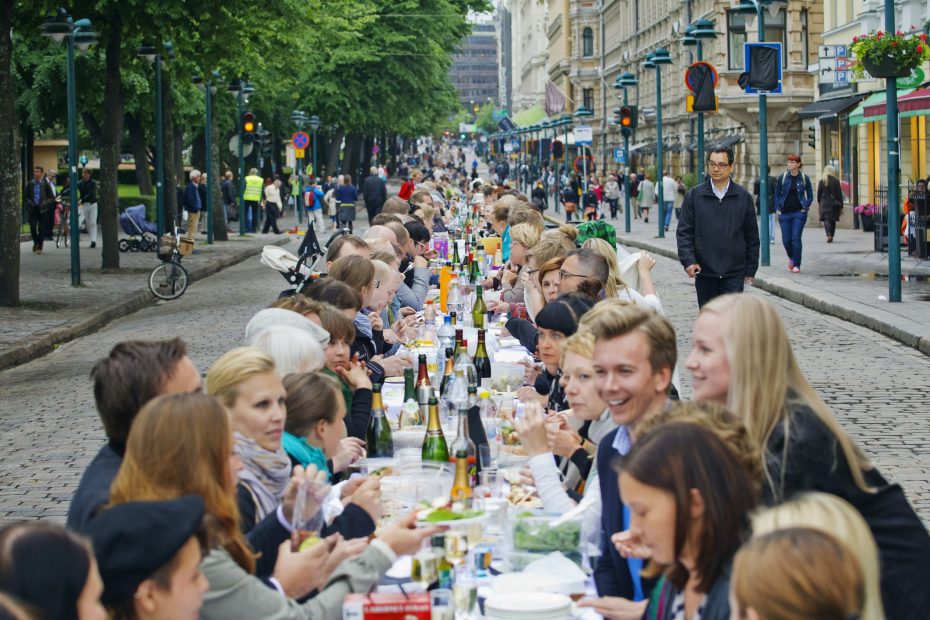 Paljon ihmisiä istuu pitkän ruokapöydän ääressä syömässä keskellä kaupunkia.