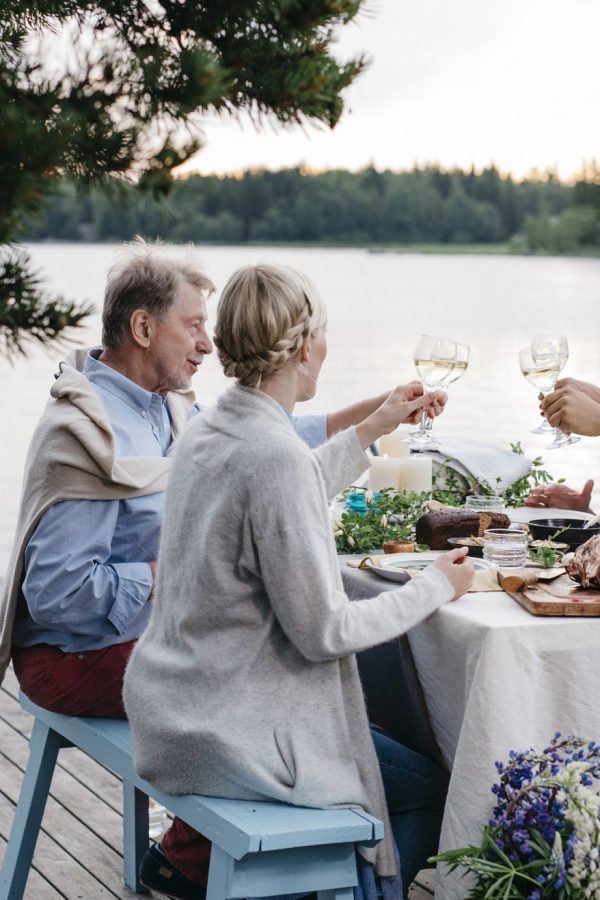 Hymyilevä pöytäseurue kohottaa kuohuviinilaseja järvimaisemassa.